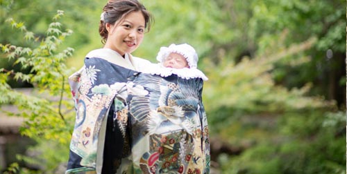 訪問着姿の母親が祝い着をまとった赤ちゃんを抱くお宮参りの様子