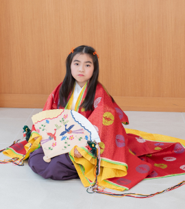 扇子を手に持ち、座ってる赤い十二単姿の7歳女児